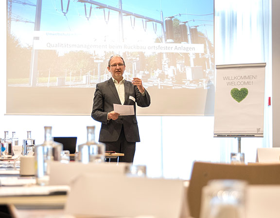 Sachverständiger Dr. Schoeps steht vor einer großen Beamerleinwand und hält einen Vortrag vor an mehreren Tischen sitzenden Kunden